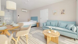 A modern hotel room with a bed, a light blue sofa, a wooden coffee table, and a dining area with a table and chairs.