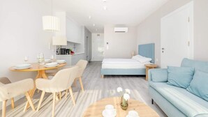 A modern hotel room with a bed, a sofa, a dining table with chairs, a coffee table, and a kitchen area in the background.