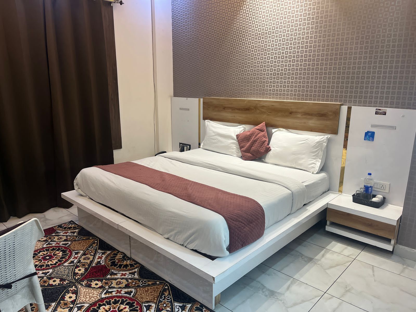Habitación de hotel con una cama grande, una mesita de noche con una botella de agua y una alfombra estampada.