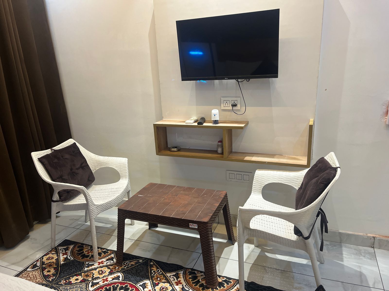 Une petite pièce avec un téléviseur fixé au mur, une étagère en bois sur laquelle se trouvent une lampe et un téléphone, deux fauteuils en osier blancs et un tapis à motifs.