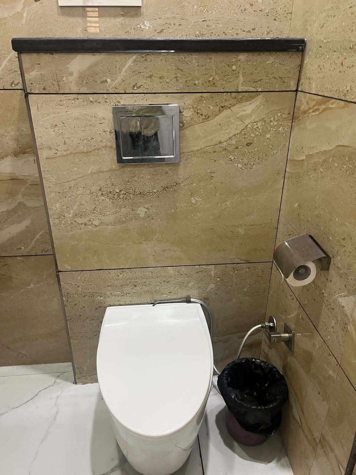 Casa de banho com sanita branca, um contentor de lixo preto e um dispensador de toalhas de papel.