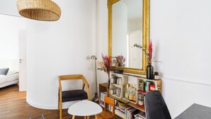 A room with a white sofa, a wooden chair, a small white table, a bookshelf with various items, and a large mirror with a gold frame.