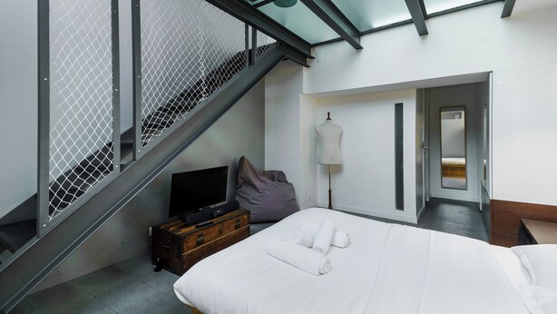 A modern hotel room with a bed, a television, a suitcase, and a staircase with a glass railing.