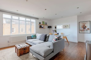 A modern living room with a grey sectional sofa, a wooden chest, and a dining area with a table and chairs. - Contemporary 2BD Flat with Balcony, Shoreditch (Greater London)
