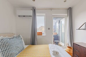 A hotel room with a bed, a dresser, a window with curtains, an air conditioning unit, and a view of buildings outside. - Contemporary 2BD Flat with Balcony, Shoreditch (Greater London)