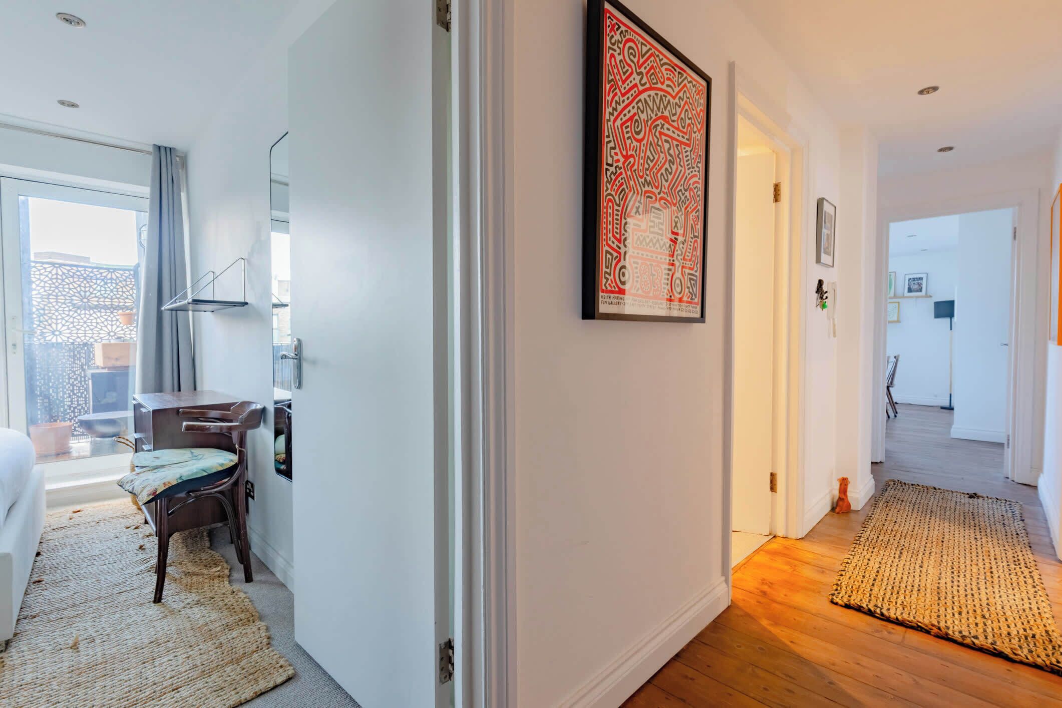 A hallway with a wooden floor, a woven rug, a framed artwork, a desk with a chair, and a bed in the background.