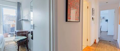 A hallway with a wooden floor, a woven rug, a framed artwork, a desk with a chair, and a bed in the background.