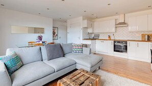 Un salon moderne avec un canapé gris, une table basse en bois et une cuisine dotée de meubles blancs et d’un four à micro-ondes.