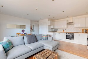 A modern living room with a grey sofa, wooden coffee table, and a kitchen with white cabinets and a microwave. - Contemporary 2BD Flat with Balcony, Shoreditch (Greater London)