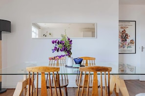 A dining area with a glass table, wooden chairs, a vase with purple flowers, a white pitcher, and a blue bowl. - Contemporary 2BD Flat with Balcony, Shoreditch (Greater London)