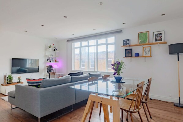 A modern living room with a glass dining table, grey sofa, and a flat-screen TV.