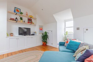 Un salon moderne avec un téléviseur à écran plat, un canapé agrémenté de coussins à motifs, une plante en pot et une bibliothèque contenant divers objets.