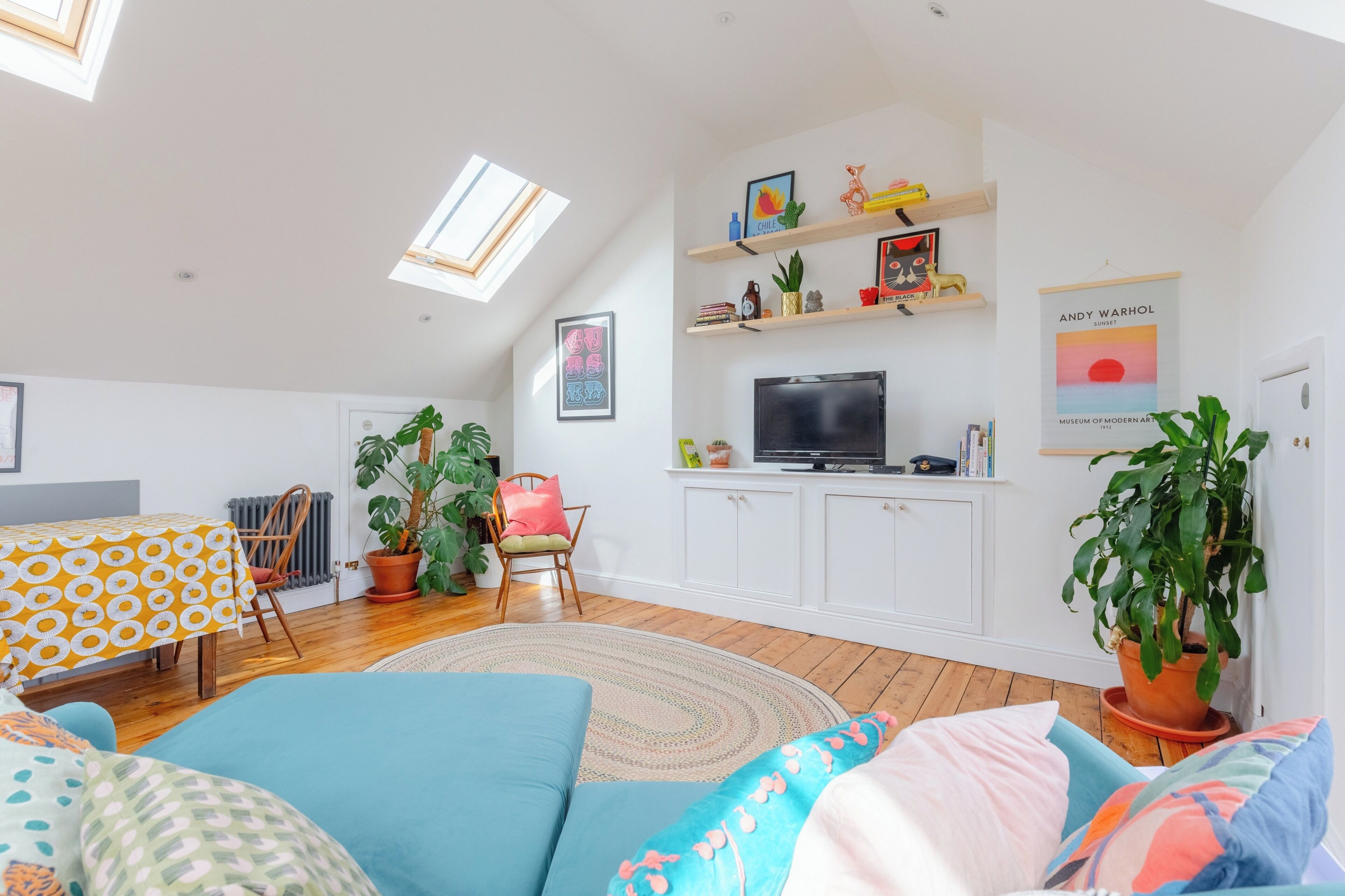 A modern living room with a sofa, a dining table, and a TV. There are plants and artwork on the walls.