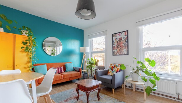 Ein Wohnzimmer mit einer Wand in Türkis, einem runden Spiegel, einer Couch, einem Couchtisch und einem Essbereich mit Stühlen.