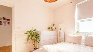 Ein Schlafzimmer mit Bett, weißen Wänden, einem Blumentopf, einem Schminktisch und einem Fenster.