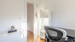 Ein modernes, minimalistisches Zimmer mit Holzboden, Schreibtisch und Stuhl, einem weißen Schminktisch und einer Tür zu einem weiteren Raum.