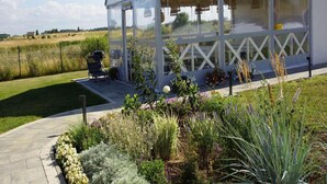 A covered outdoor area with a grill and seating, surrounded by a garden with various plants and flowers. - Comfortable Holiday Homes With air Conditioning (Mielno)