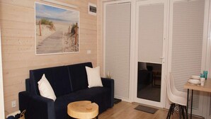A room with a dark blue sofa, a small wooden stool, a dining table with chairs, and a large window with blinds. - Comfortable Holiday Homes With air Conditioning (Mielno)