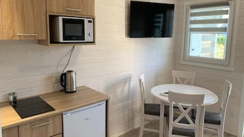 Cozinha compacta com micro-ondas, chaleira e pequena geladeira. Há uma área de jantar com mesa e cadeiras, e uma TV fixada na parede.