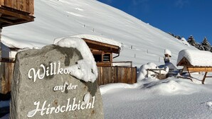 Ein Steinschild mit der Aufschrift 'Willko auf der Hirschbichl Alm', Holzbauten und verschneite Landschaft.
