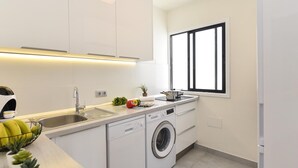 Una cucina moderna con mobili bianchi, lavastoviglie integrata, lavatrice e una finestra con infissi neri.