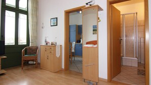 Una habitación con piso de madera, una silla de mimbre, un armario de madera, una mesita con una lámpara y una puerta que da a un baño con ducha.