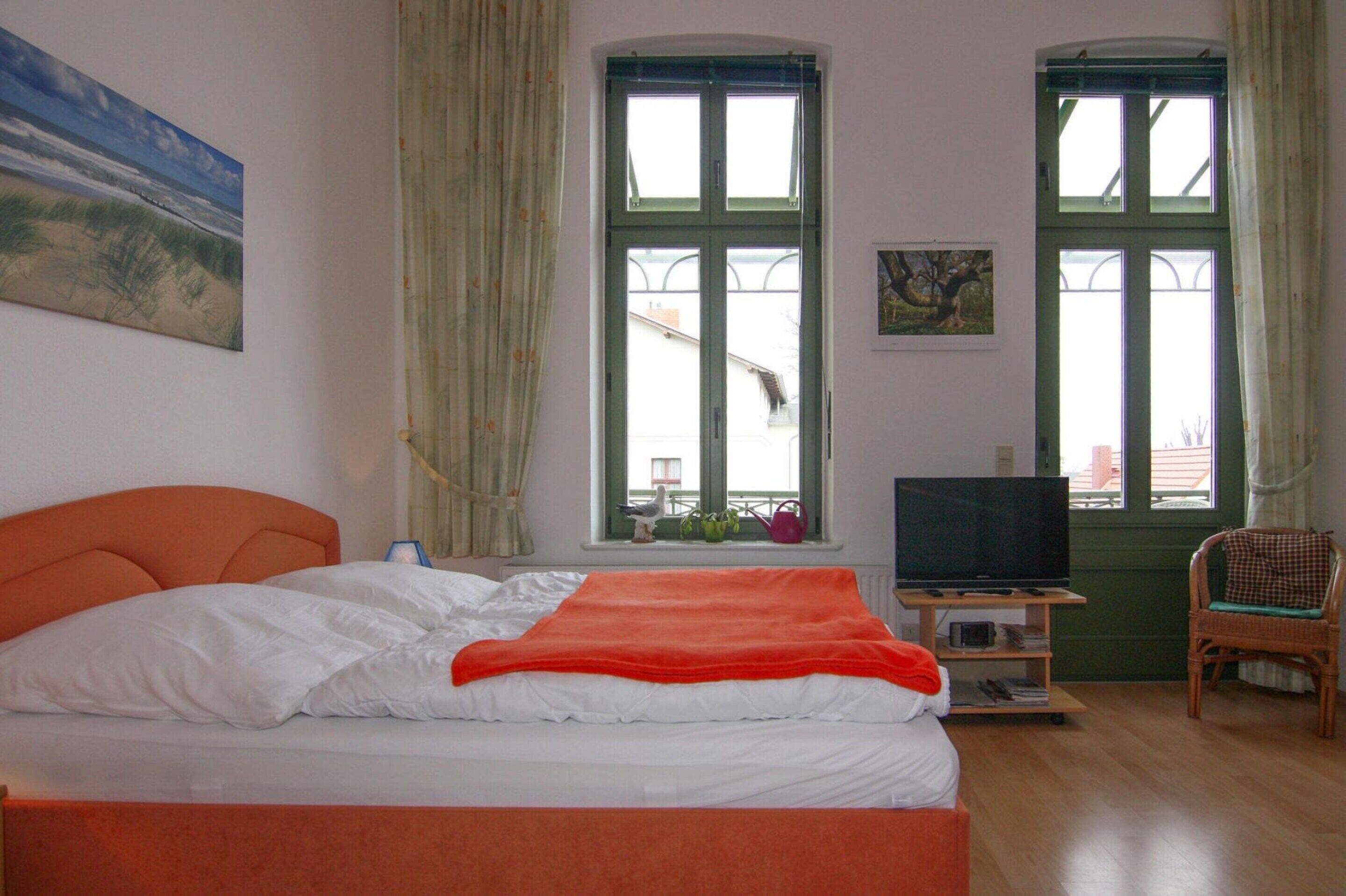 Ein Schlafzimmer mit einem Bett, zwei Fenstern, einem Fernseher und einem Sessel.