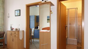 Una habitación con un armario de madera, una silla, una mesita con lámpara y un espacio con ducha.