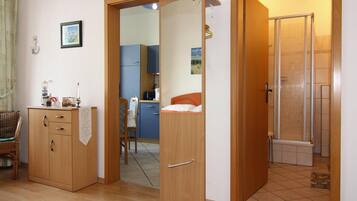 A room with a wooden wardrobe, a chair, a small table with a lamp, and a shower area.