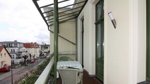 Un balcón con una silla y una mesa de mimbre, con vista a una calle con coches aparcados y edificios.