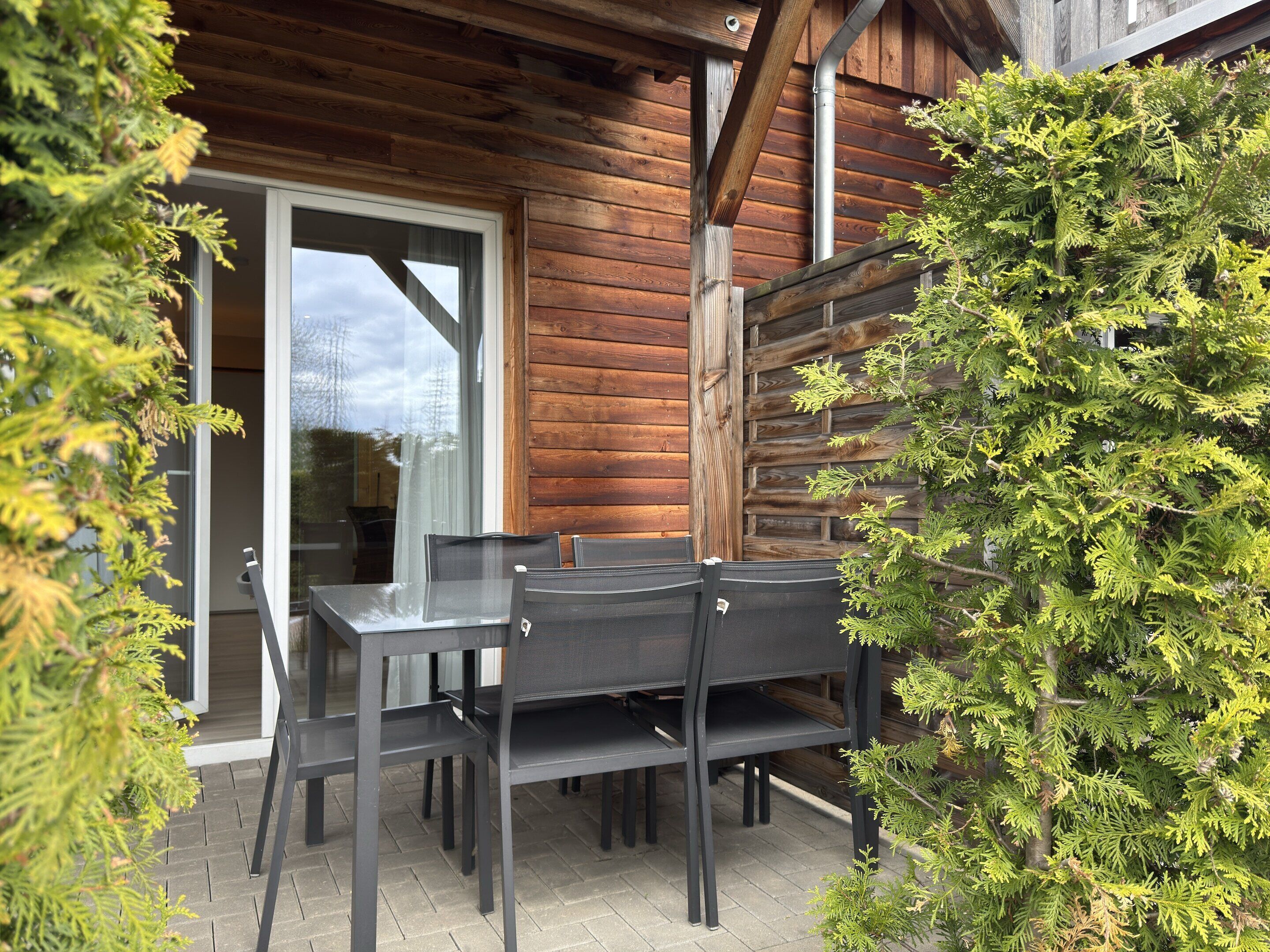 An outdoor patio with a wooden table and chairs, surrounded by greenery.