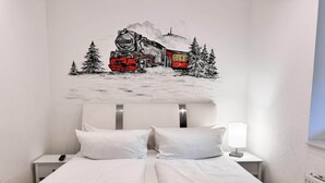 Een slaapkamer met een bed, wit beddengoed, een nachtkastje met een lamp en een decoratie van een trein aan de muur.