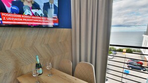 A hotel room with a wooden table, two wine glasses, and a bottle of wine. A television is mounted on the wall displaying a news broadcast. The room has a view of the sea and parked cars outside. - Comfortable Apartment With a sea View (Ustronie Morskie)