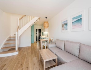 A modern living room with a wooden staircase, a light-colored sofa, a wooden coffee table, and a dining area with a table and chairs. - Holiday House for 5 People in Grzybowo (Kolobrzeg)