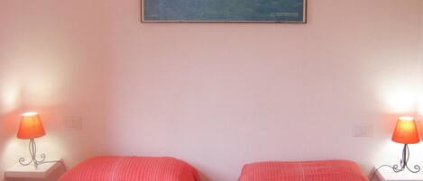 A hotel room with two single beds, each with a red-striped bedspread, two bedside tables with lamps, and a framed picture of a coastal scene on the wall.