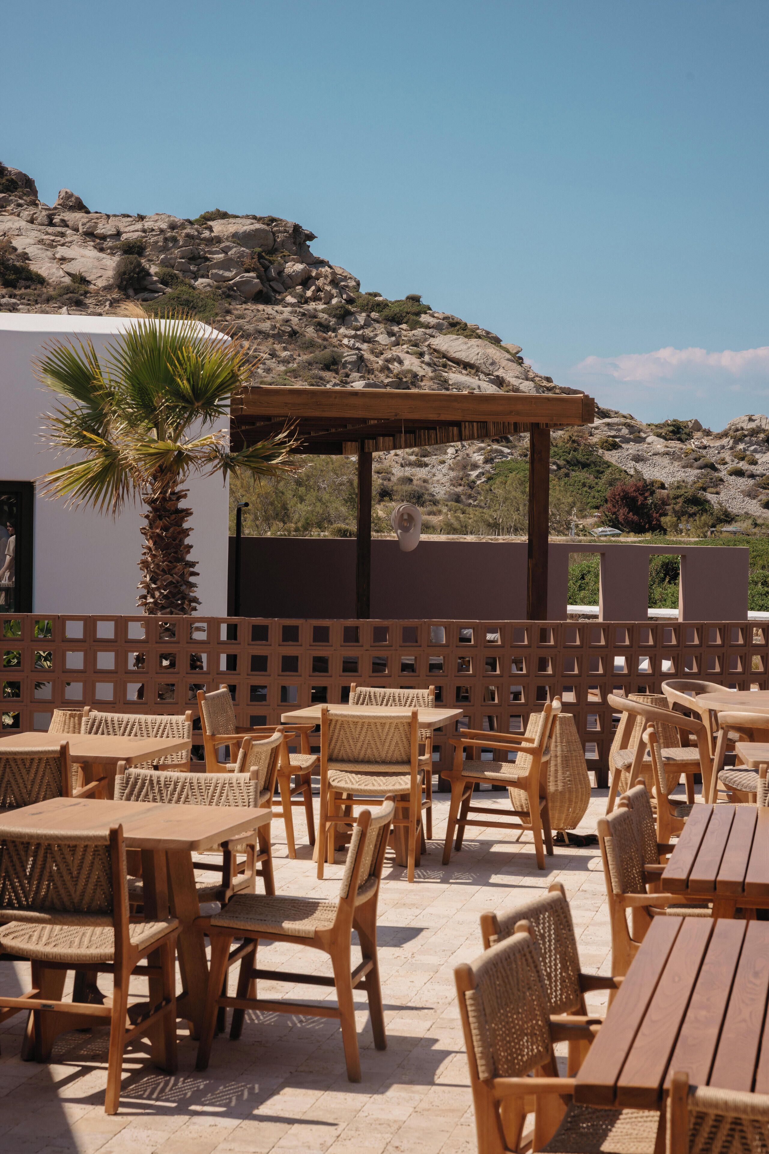 Un patio exterior con mesas y sillas de madera, una palmera y vistas a colinas rocosas.
