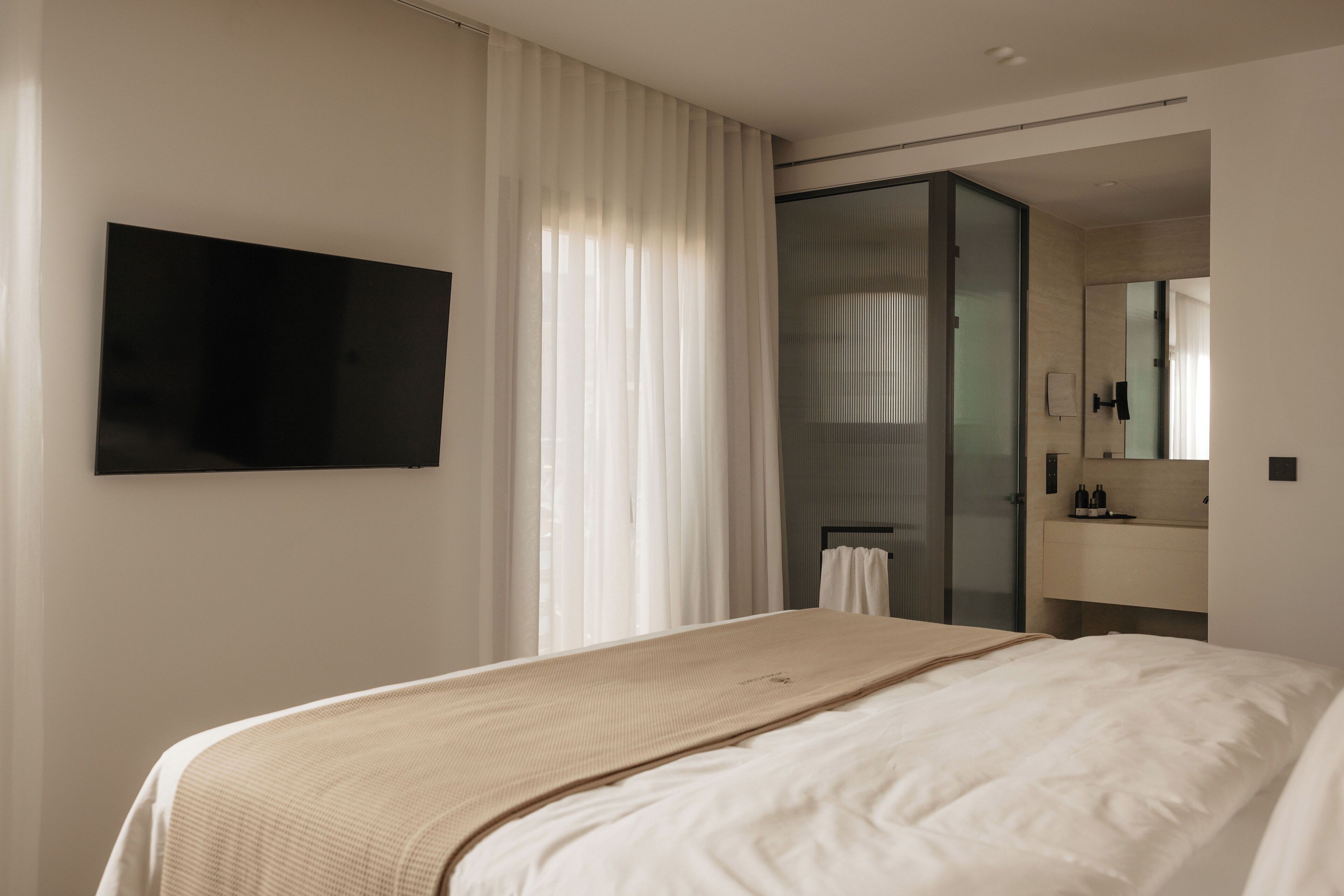 A modern hotel room with a large bed, a flat-screen TV mounted on the wall, a bathroom with a glass shower enclosure, and a small sink with a mirror.