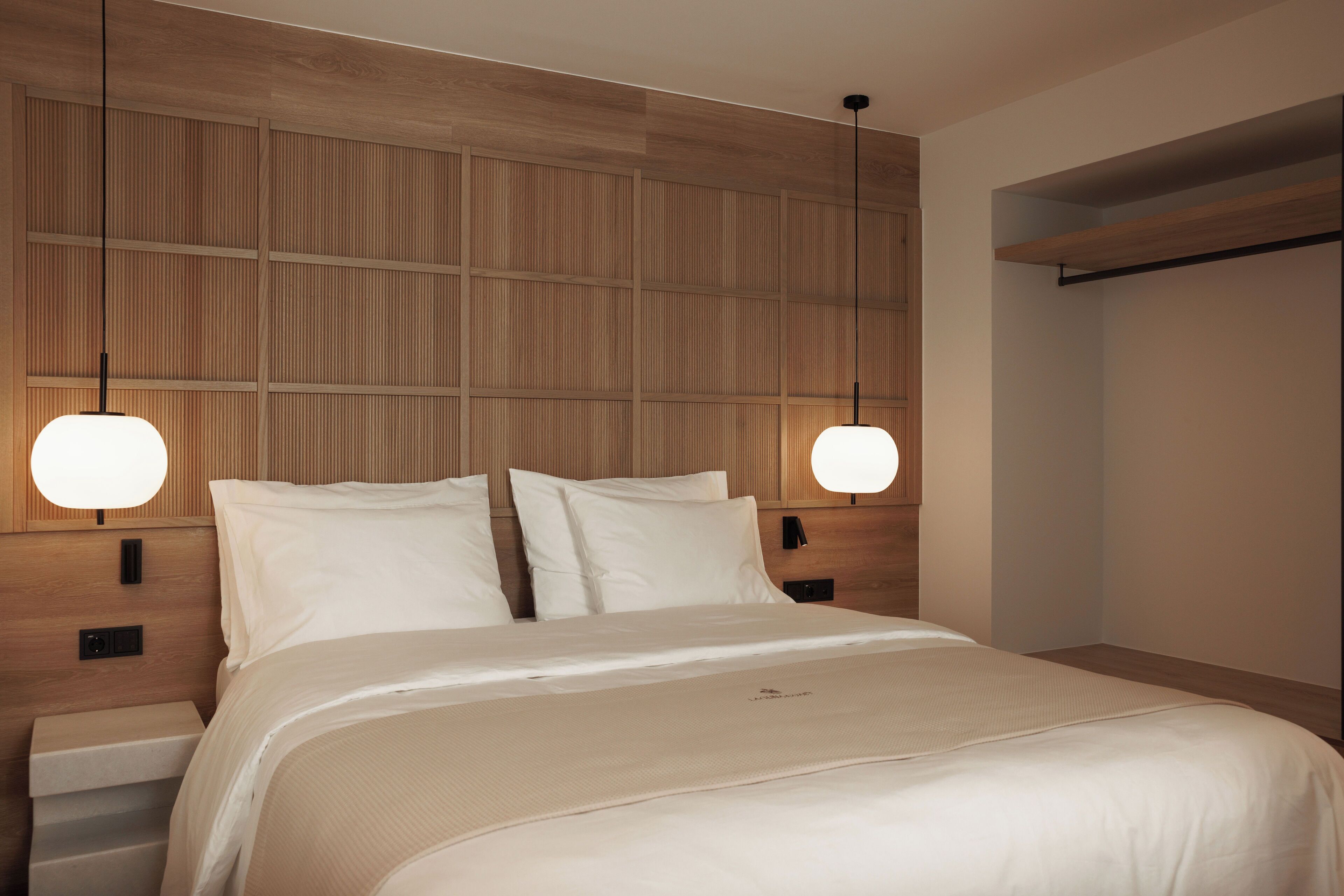 A modern hotel room with a large bed, wooden headboard, and pendant lighting.