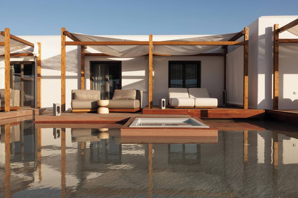A modern outdoor pool area with wooden pergolas, lounge chairs, and a clear pool reflecting the surroundings.