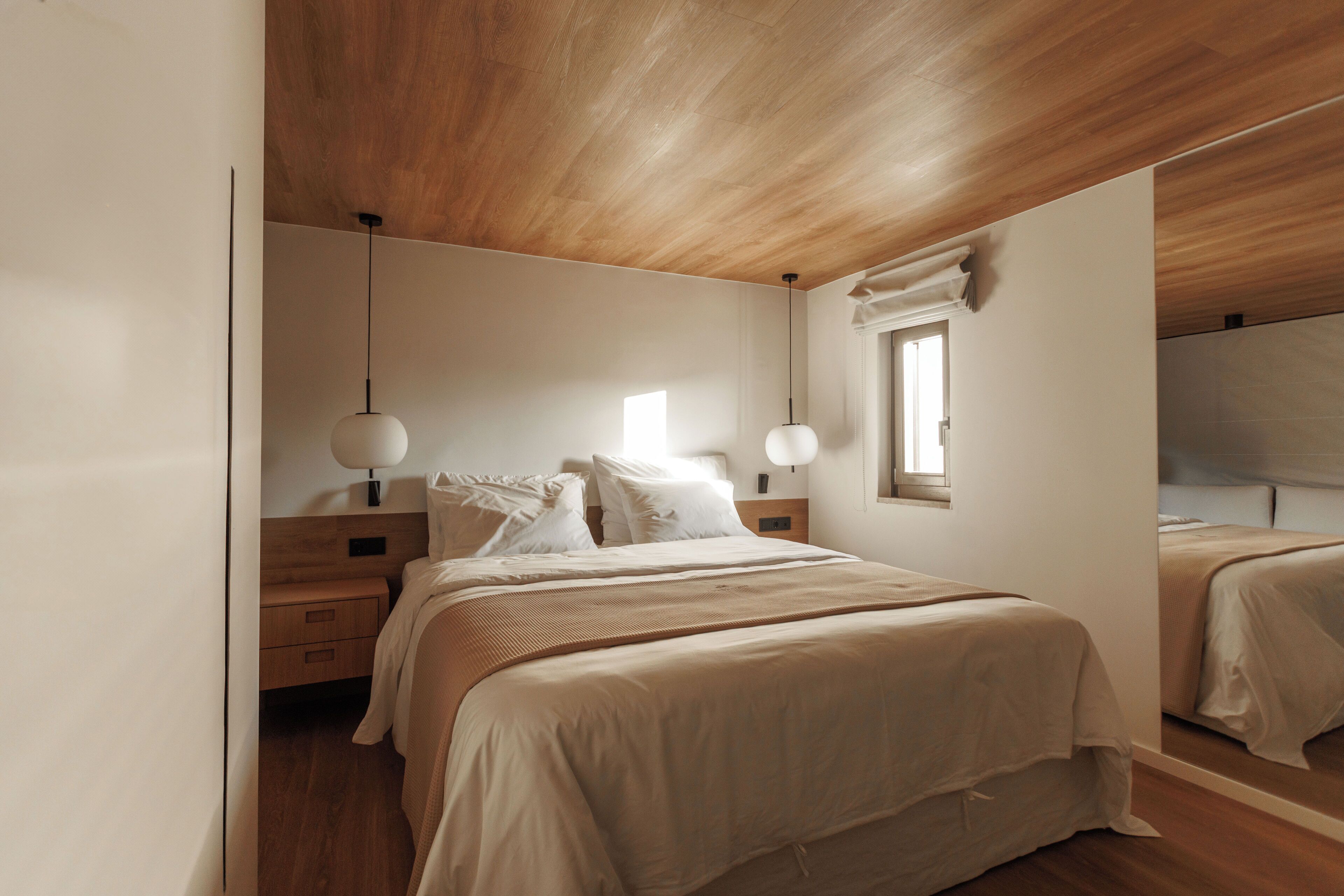 Ein Schlafzimmer mit Holzbalkendecke, einem großen Bett mit weißer Bettwäsche, zwei Nachttischen und einem Fenster mit Tageslicht.