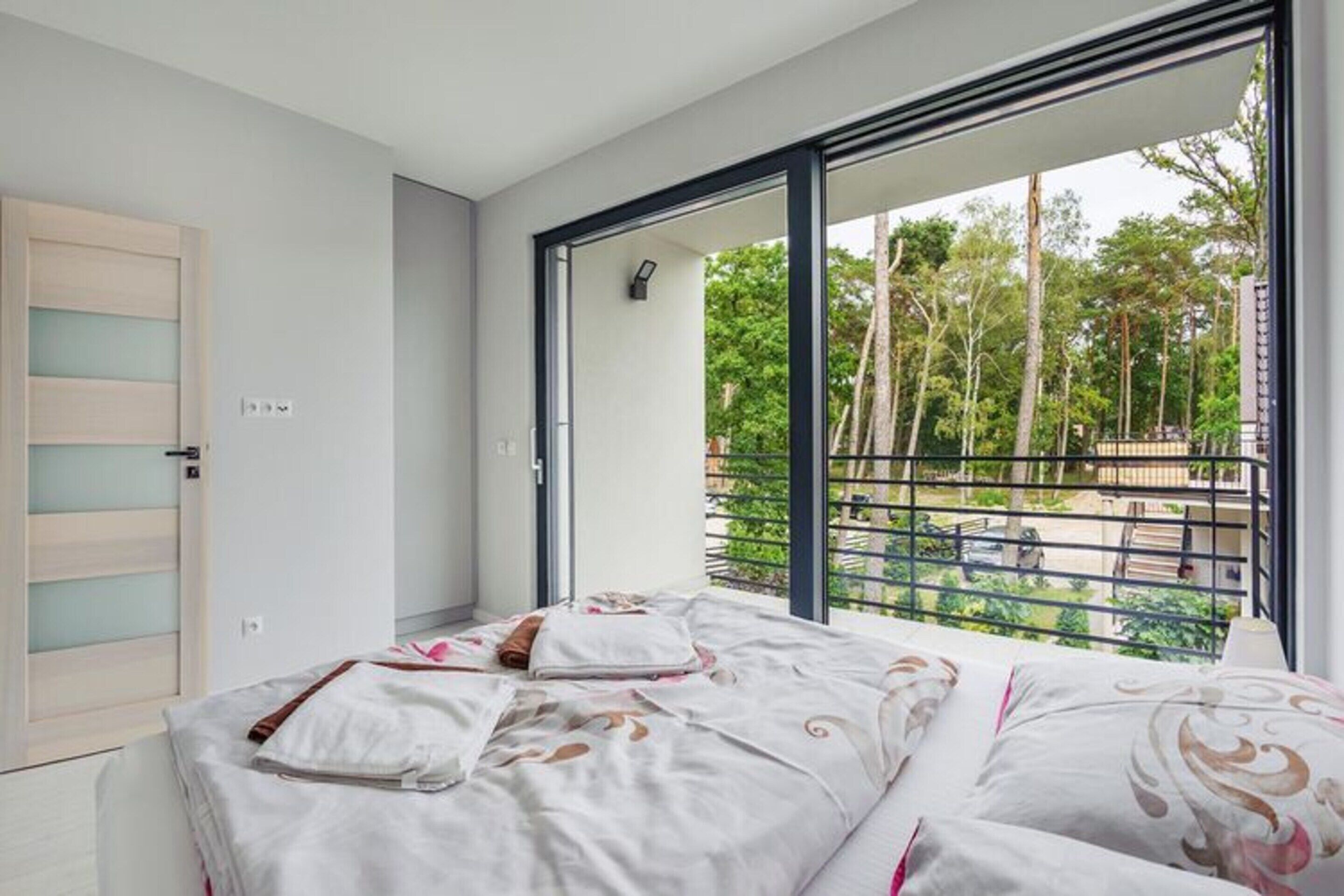 A bedroom with a large window, a bed with patterned bedding, and a view of a balcony with greenery.