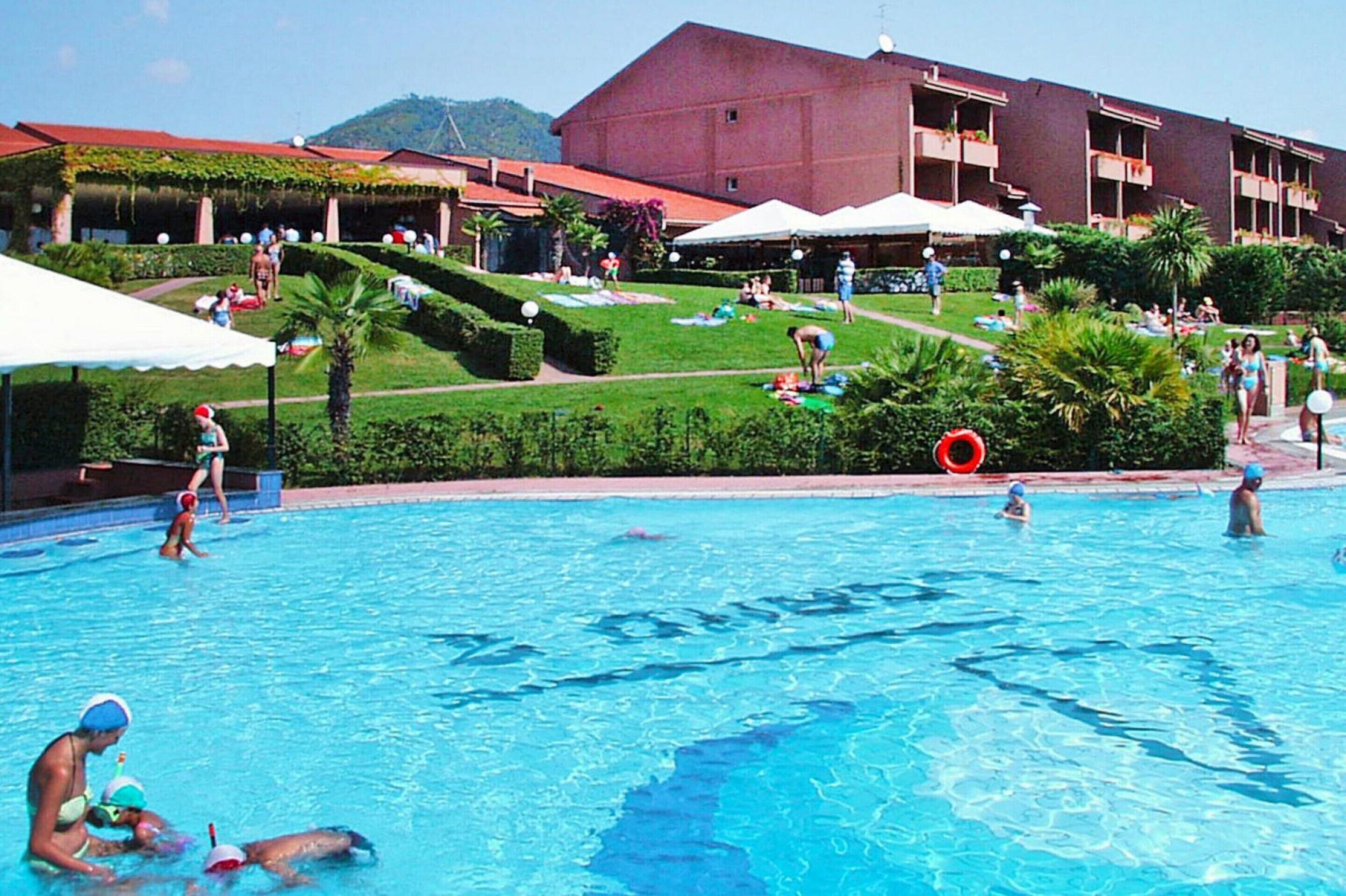 A swimming pool area with people swimming and sunbathing, surrounded by well-maintained lawns and a large building in the background.