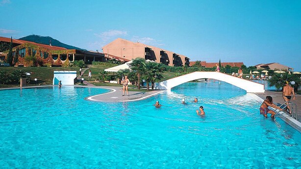 Una gran piscina al aire libre con un puente blanco, palmeras y edificios al fondo.
