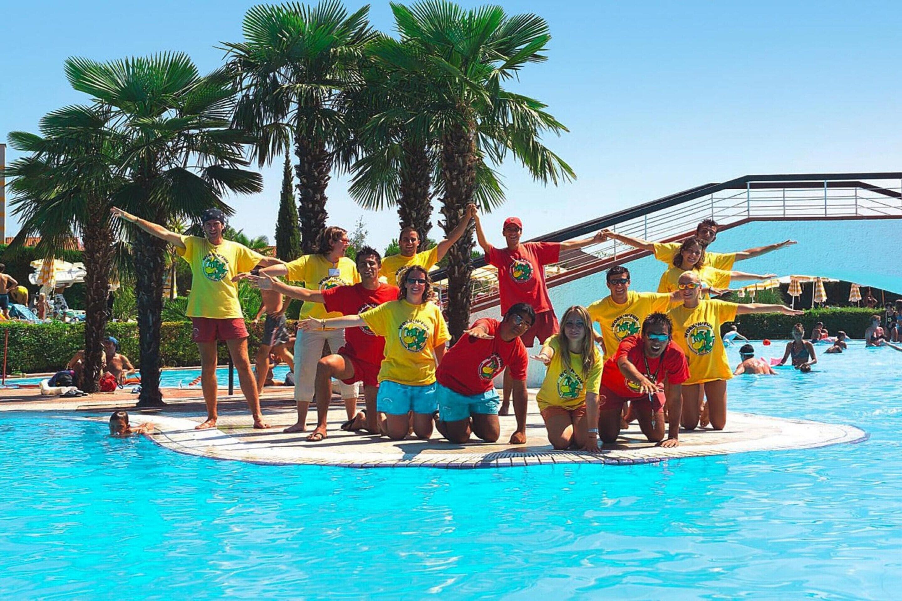 A group of people in yellow and red shirts posing by a pool with palm trees and a clear blue sky in the background.