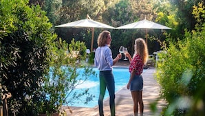 Twee personen staan bij een zwembad met parasols, omringd door groen.
