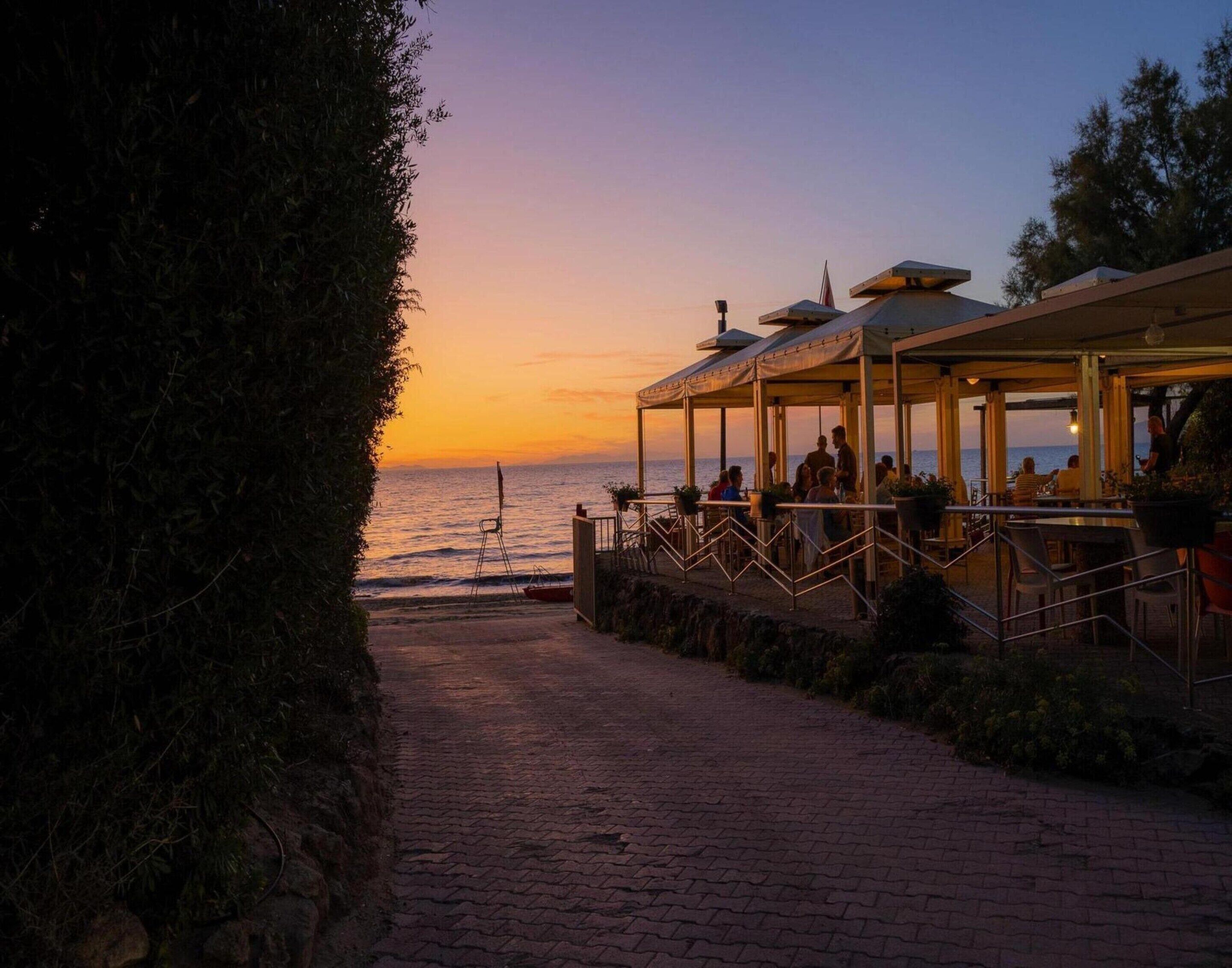 Un restaurante frente al mar con terraza al aire libre, un camino pavimentado y vista al atardecer.