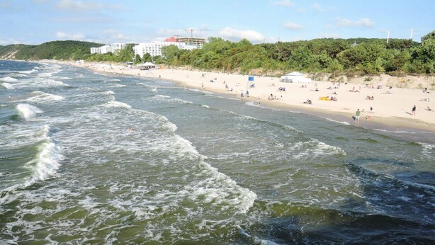 Ein Strand mit Wellen, sandigem Ufer und Gebäuden im Hintergrund.