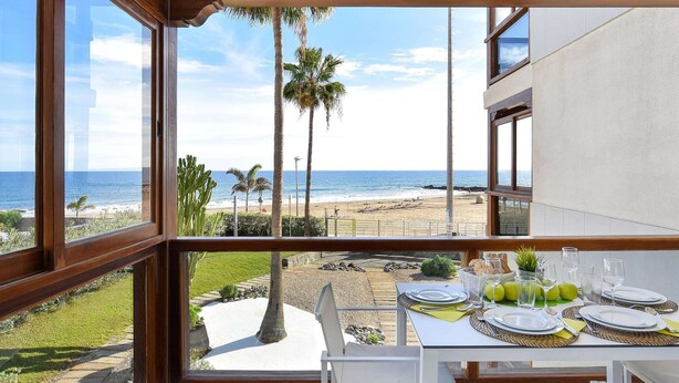 A dining area with a table set for four, overlooking a beach with palm trees and the ocean. - Apartment, San Agustin (San Bartolomé de Tirajana)