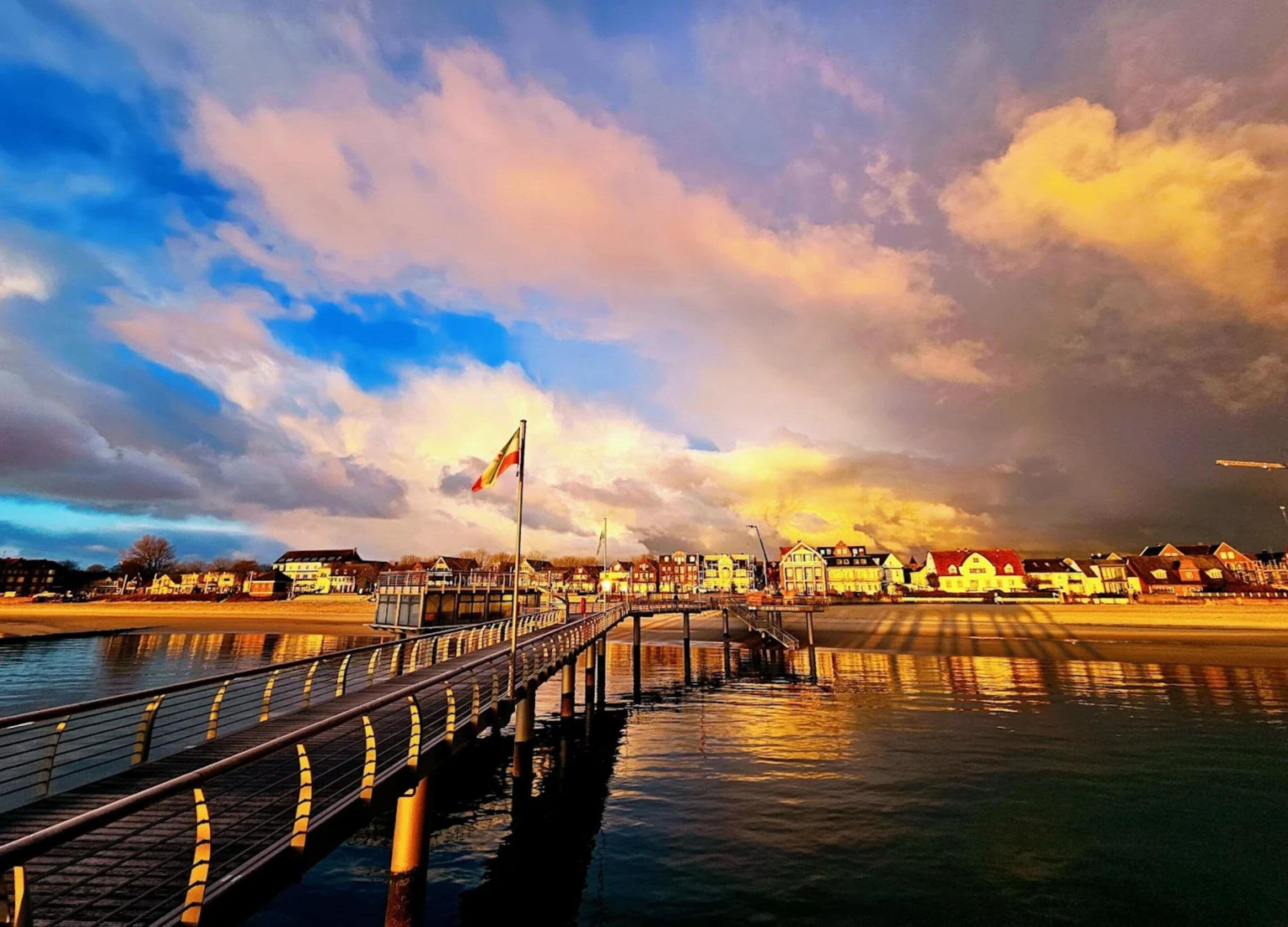 En strandpromenad vid vattnet med en bro, en flagga och byggnader i bakgrunden.