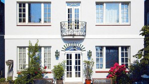 A two-story building with a sign reading "GUD HOOR". There are two windows on each floor, a balcony with ornate railings, and a small garden area with benches and potted plants. - Ferienwohnung, Wyk auf Fohr (Wyk auf Foehr)